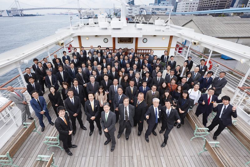 客船のデッキに並んだ社員の集合写真。背景に東京湾とレインボーブリッジ