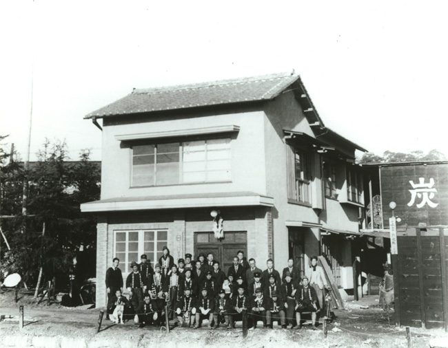 「炭」の看板を掲げた木造2階建ての建物と、その前に並ぶ人びとの集合写真
